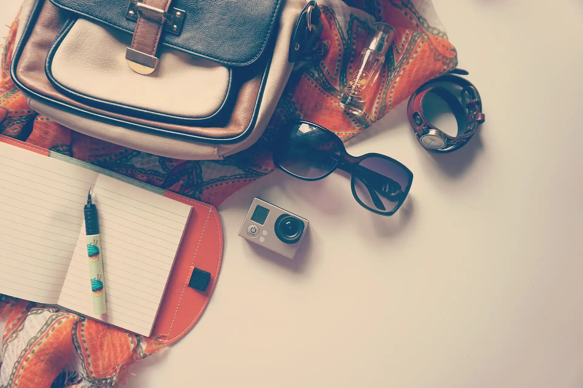 Nell'immagine, un primo piano su un tavolo sul quale sono poggiate un foulard, una borsa, un bloc-notes con una penna, una macchinetta fotografica, un paio di occhiali da sole e un orologio.