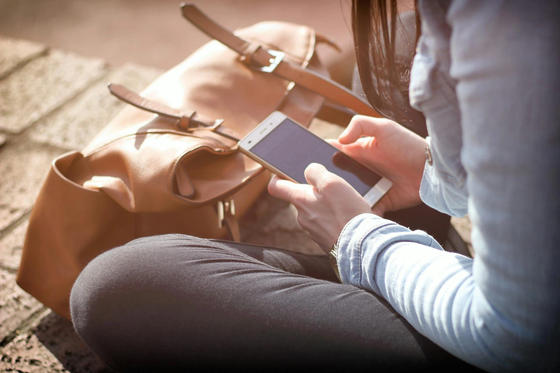 Nell'immagine, il primo piano sulle mani di una ragazza che sta guardando il cellulare, seduta a gambe incrociate. Davanti a lei è poggiata la sua borsa.